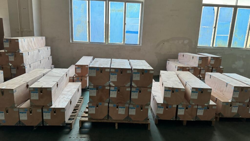 Warehouse storage area filled with stacks of boxed electric fireplaces. Multiple pallets hold rows of large cardboard boxes, each labeled with product information. The room features bare concrete walls and blue-tinted windows, suggesting an industrial setting. This image showcases the scale of electric fireplace production and distribution, highlighting the growing popularity of these modern heating solutions.